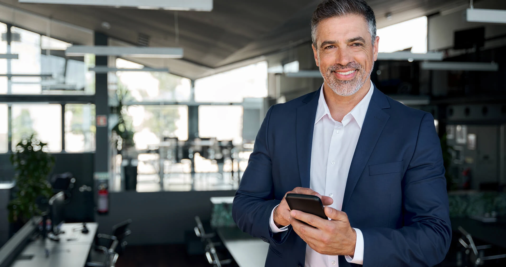 Ein Mann mittleren Alters steht in einem modernen, hellen Büro. Er trägt einen dunkelblauen Anzug und ein weißes Hemd, hält ein Smartphone in den Händen und lächelt freundlich in die Kamera. Im Hintergrund sind große Fenster und Arbeitsplätze zu sehen.
