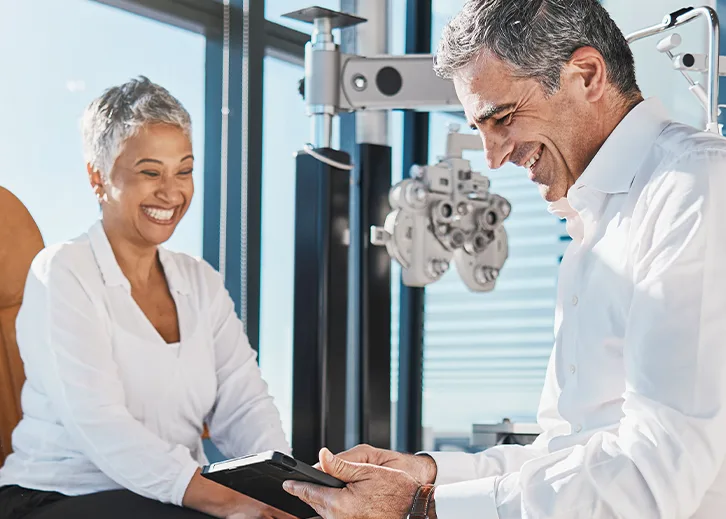 A woman and a doctor are visible. They are discussing options, i.e., a consultation is taking place. Eye examination equipment can be seen in the background.